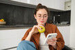Young woman chilling at home, looking at smartphone, reading news on social media and eating green apple in the kitchen