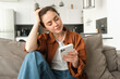 Portrait of beautiful young woman reading news on mobile phone, using smartphone app, looking at screen and smiling, sitting at home on sofa