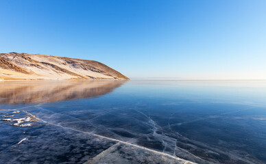 Wall Mural - Baikal Lake on cold sunny day in January. Snow-covered coastal hills are reflected in expanses of blue mirror ice with cracks at sunset. Winter beautiful landscape. Outdoor recreation and ice travel