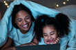 Mom, child and tablet with blanket in bedroom at night to play games, social media and reading ebook. Happy mother, girl and kid relax with digital technology, watching cartoon or movies in dark fort