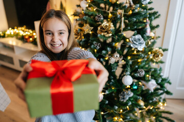 Closeup face of happy adorable little girl holding, giving and showing at camera wrapped with red ribbon and bow festive Christmas present gift box, sitting by glowing xmas tree, looking at camera