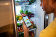© Marko Geber - Man checking a meal planning app while looking into a refrigerator filled with fresh vegetables