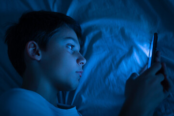 Boy using a smartphone at night in bed. 10 year old boy playing with mobile phone before going to sleep. Concept of children using the smartphone in their room alone without control. Copy space