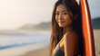 A young, smiling Chinese woman stands on the beach with her surfboard, her skin beautifully bronzed by the sun.