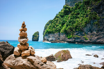 Wall Mural - Zen rock tower stands at the forefront of a breathtaking tropical coastline. Behind, emerald waves crash against rugged cliffs adorned with green foliage.