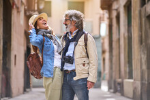 Laughing Adult Caucasian Couple Standing Embraced Affectionately In Tourist Street Of Town. Mature People In Love Enjoying Romantic Vacation Time. Positive Relationships And Weekend Getaways Spring.