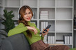© wichayada - Happy Asian woman listening to music on mobile phone while sitting on sofa at home Relaxed smiling girl with headphones in the morning It's time to rest.