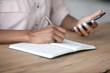 © fizkes - Hands of student girl watching learning webinar on smartphone, writing in notebook, preparing for exams. Woman taking notes, planning work tasks, using online organizer app. Close up