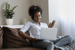 © fizkes - Happy African student girl in headphones making video call from home, using laptop, watching webinar, attending online conference, virtual class, waving hello, speaking at webcam, resting on couch