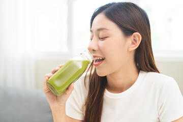 Green detox juice concept, Beautiful smile asian young woman hand holding vegetable smoothie bottle for diet at home. Happy girl drinking healthy meal food for weight loss. Lifestyle, vegan nutrition.