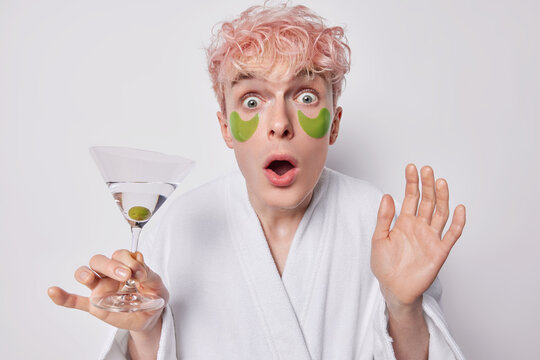 Photo of shocked terrified pink haired man applies green beauty patches under eyes to reduce puffiness holds glass of cocktail stares with omg expression at camera isolated over white background