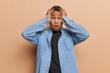 Wall Mural - Indoor waist up of young confused African american male wearing denim shirt and black t shirt standing in centre isolated on beige background not knowing what to do keeping hands raised on head