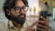 Indian male bearded man in glasses professional hold modern smartphone texting message in office indoors. Arabian businessman using mobile app browsing phone business chatting scrolling web online