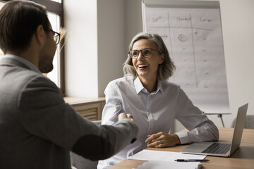 Poster - Cheerful mature business professional woman shaking hands with younger coworker man at laptop, smiling, laughing. Project partners giving greeting handshake after successful meeting