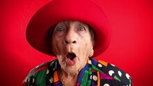 Saying WOW, a happy fisheye portrait caricature of funny elderly woman with red hat isolated on red background.