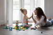 © fizkes - Happy mother playing wooden blocks with daughter, finish build tower, praising kid, giving high five settled down together on warm floor in living room. Playtime, help, happy motherhood, leisure games