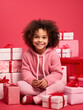 Little girl in Santa hat with lots of Christmas gifts, child wearing pink sweater sitting on the floor with lots of gifts, African-American girl smiling looking at camera, pink background