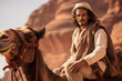 Side view of man in traditional Saudi dish dash, kaffiyeh, and agal sitting astride dromedary camel with red sandstone cliffs in background