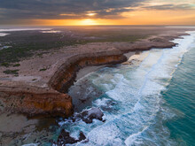 Aerial View Of Waves Crashing Against Eroded Cliffs Along A Rugged Coastline At Sunrise
