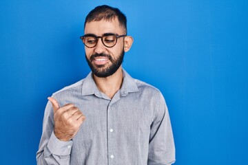 Wall Mural - Middle east man with beard standing over blue background smiling with happy face looking and pointing to the side with thumb up.