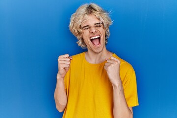 Poster - Young modern man standing over blue background excited for success with arms raised and eyes closed celebrating victory smiling. winner concept.