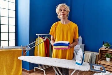 Poster - Young man ironing clothes at home making fish face with mouth and squinting eyes, crazy and comical.