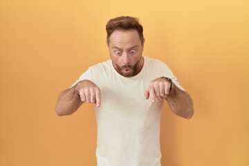 Wall Mural - Middle age man with beard standing over yellow background pointing down with fingers showing advertisement, surprised face and open mouth