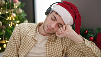 Poster - Young hispanic man sleeping on sofa by christmas tree wearing headphones at home