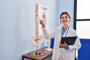 Canvas Print - Young blonde woman pysiotherapist pointing to anatomical model of vertebral column holding clipboard at rehab clinic