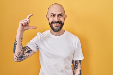 Poster - Young hispanic man with tattoos standing over yellow background smiling and confident gesturing with hand doing small size sign with fingers looking and the camera. measure concept.