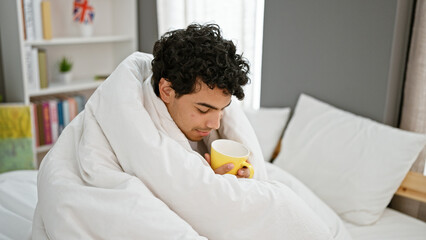 Poster - Young latin man smelling cup of coffee covering with blanket at bedroom