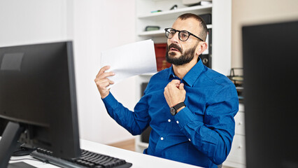 Young hispanic man business worker using computer using documents as a hand fan at office