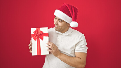 Wall Mural - Young hispanic man smiling confident holding christmas present over isolated red background