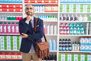 Poster - Hispanic man with beard working as salesman at pharmacy drugstore serious face thinking about question with hand on chin, thoughtful about confusing idea