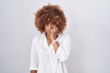 Sticker - Young hispanic woman with curly hair standing over white background looking stressed and nervous with hands on mouth biting nails. anxiety problem.