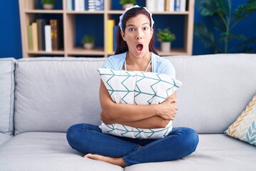 Wall Mural - Young hispanic woman hugging pillow sitting on the sofa afraid and shocked with surprise and amazed expression, fear and excited face.