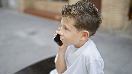 Wall Mural - Adorable blond boy engrossed in serious phone talk, sitting on city bench, relaxed amidst urban outdoor backdrop, using communication technology.