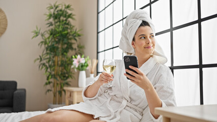 Poster - Young beautiful hispanic woman wearing bathrobe drinking champagne using smartphone at bedroom