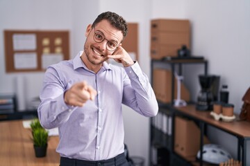 Canvas Print - Young hispanic man at the office smiling doing talking on the telephone gesture and pointing to you. call me.