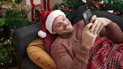 Wall Mural - Young caucasian man using smartphone lying on the sofa by christmas tree at home