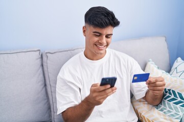 Wall Mural - Young hispanic man using smartphone and credit card sitting on sofa at home