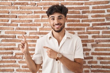 Canvas Print - Arab man with beard standing over bricks wall background smiling and looking at the camera pointing with two hands and fingers to the side.