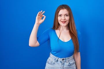 Sticker - Redhead woman standing over blue background smiling positive doing ok sign with hand and fingers. successful expression.