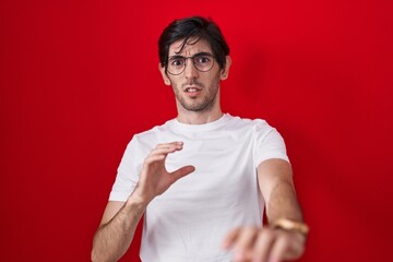 Poster - Young hispanic man standing over red background disgusted expression, displeased and fearful doing disgust face because aversion reaction.