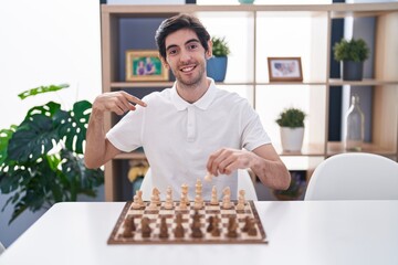 Poster - Young hispanic man playing chess sitting on the table pointing finger to one self smiling happy and proud