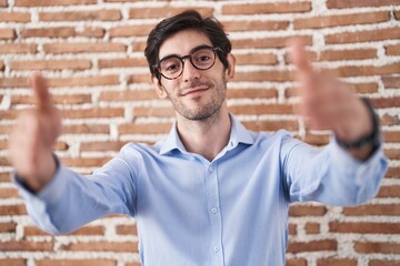 Poster - Young hispanic man standing over brick wall background looking at the camera smiling with open arms for hug. cheerful expression embracing happiness.