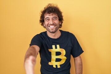 Canvas Print - Hispanic young man wearing bitcoin t shirt smiling friendly offering handshake as greeting and welcoming. successful business.