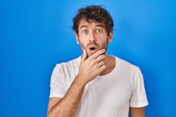 Poster - Hispanic young man standing over blue background looking fascinated with disbelief, surprise and amazed expression with hands on chin