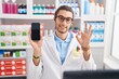 © Krakenimages.com - Young hispanic man working at pharmacy drugstore showing smartphone screen doing ok sign with fingers, smiling friendly gesturing excellent symbol
