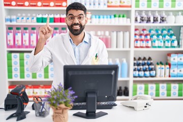 Wall Mural - Hispanic man with beard working at pharmacy drugstore showing and pointing up with finger number one while smiling confident and happy.